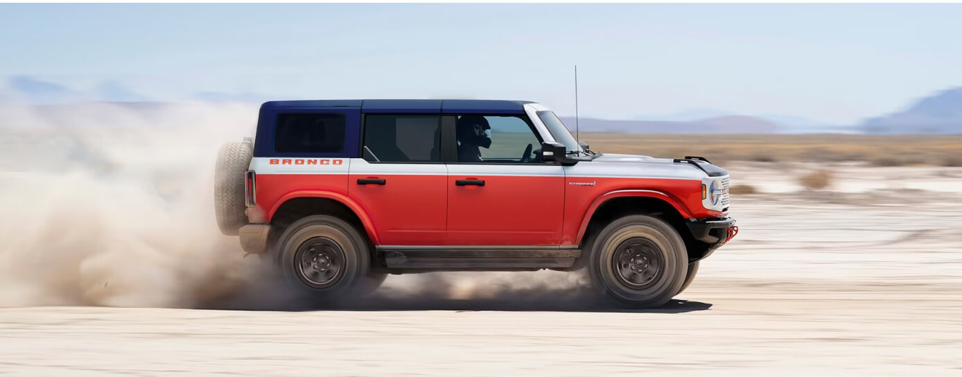 A red white and blue Ford Bronco drives down a sandy trail.