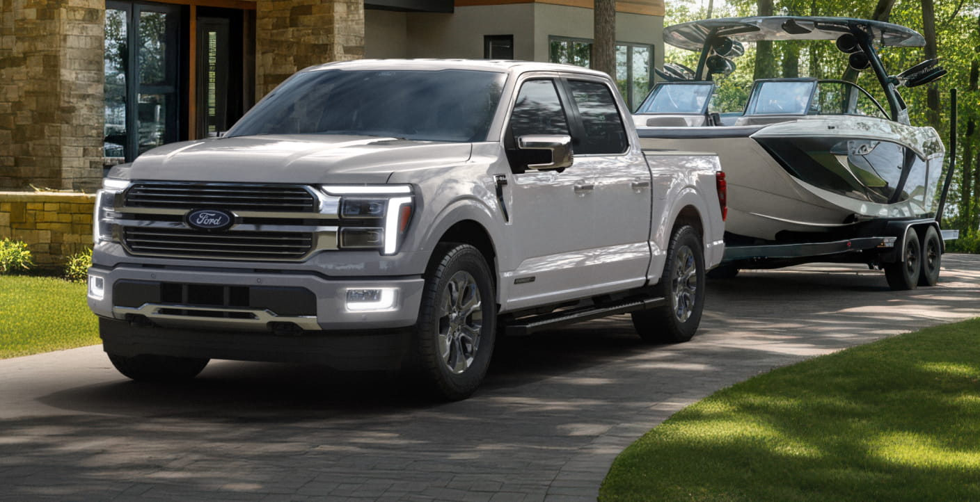 White Ford F-150 PowerBoost towing a boat on a trailer outside a modern home, showcasing the truck's towing capability.