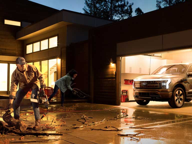 A man and woman cleaning up their driveway from a storm while their F-150 Lightning is charging the house.
