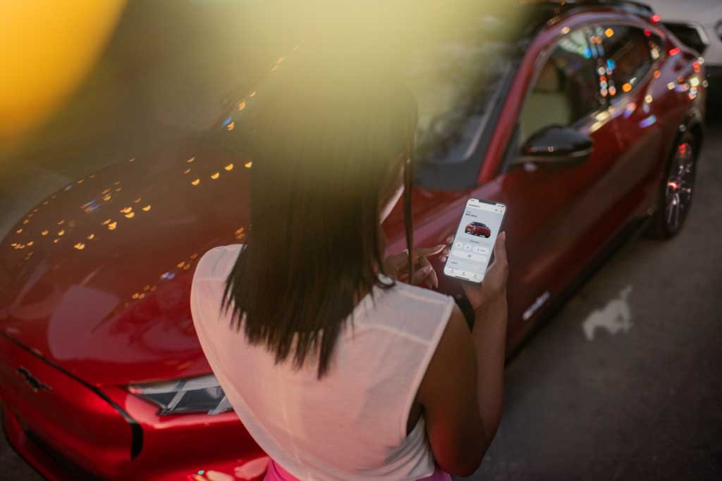 Woman using the FordPass® App on a phone while standing beside a Ford Mustang Mach-E® SUV
