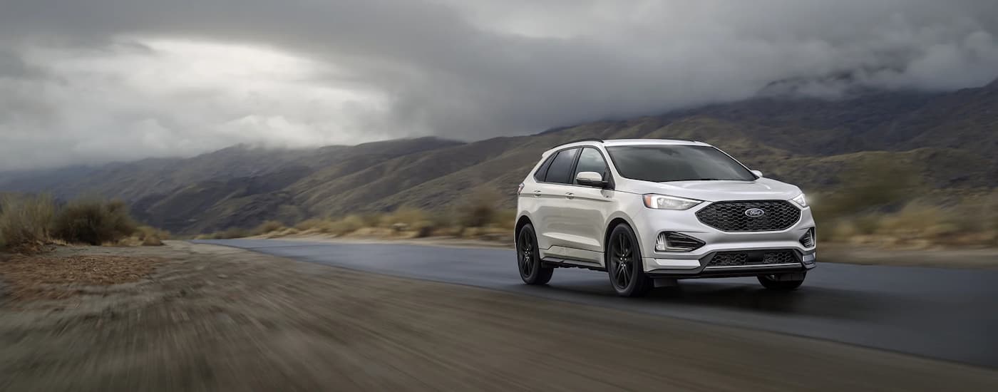 A white 2022 Ford Edge ST is shown driving on a rural road on a cloudy day.