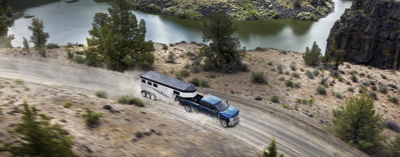 A blue 2023 Ford F-250 is shown towing a white enclosed trailer on a winding dusty road.