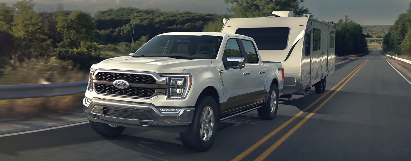 A white 2023 Ford F-150 is shown towing a camper trailer on an open highway.