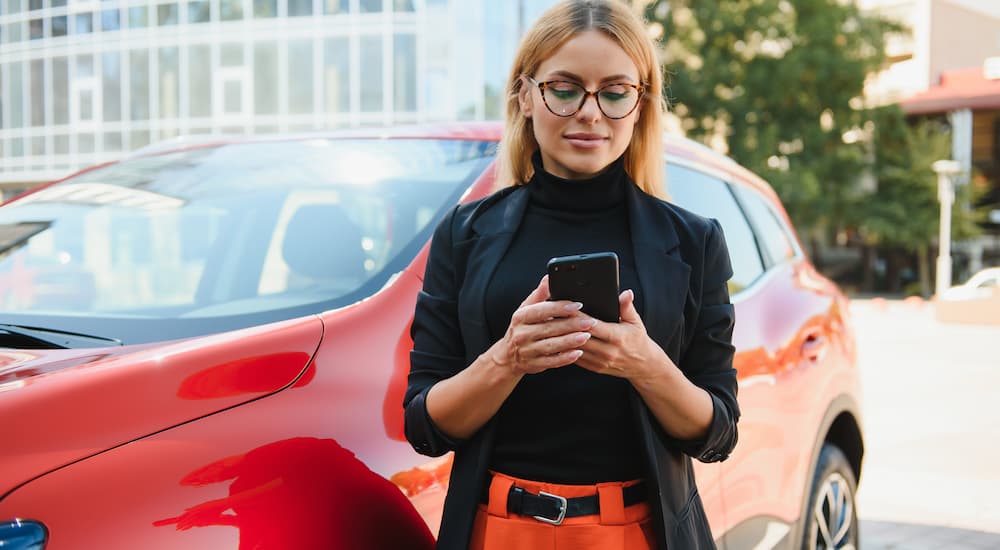 A woman is shown standing next to a red vehicle while using a cellphone.