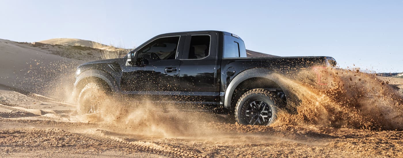 A black 2014 Ford F-150 Raptor is show from the side kicking up sand.