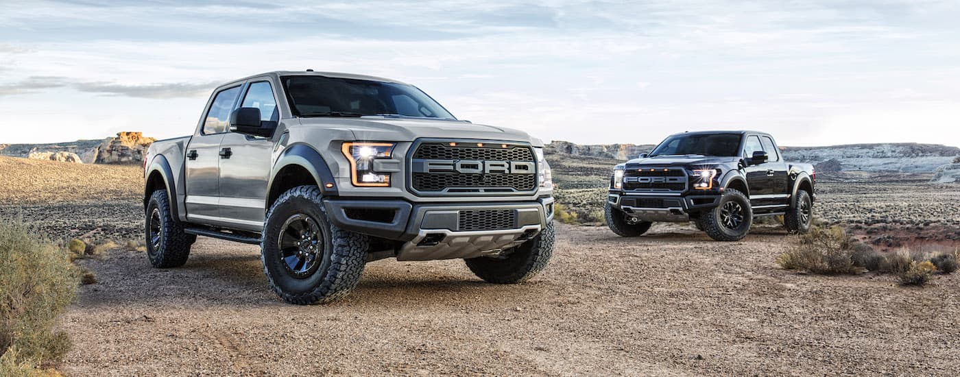 A grey and a black 2017 Ford F-150 Raptor are parking facing each other on a dirt lot.