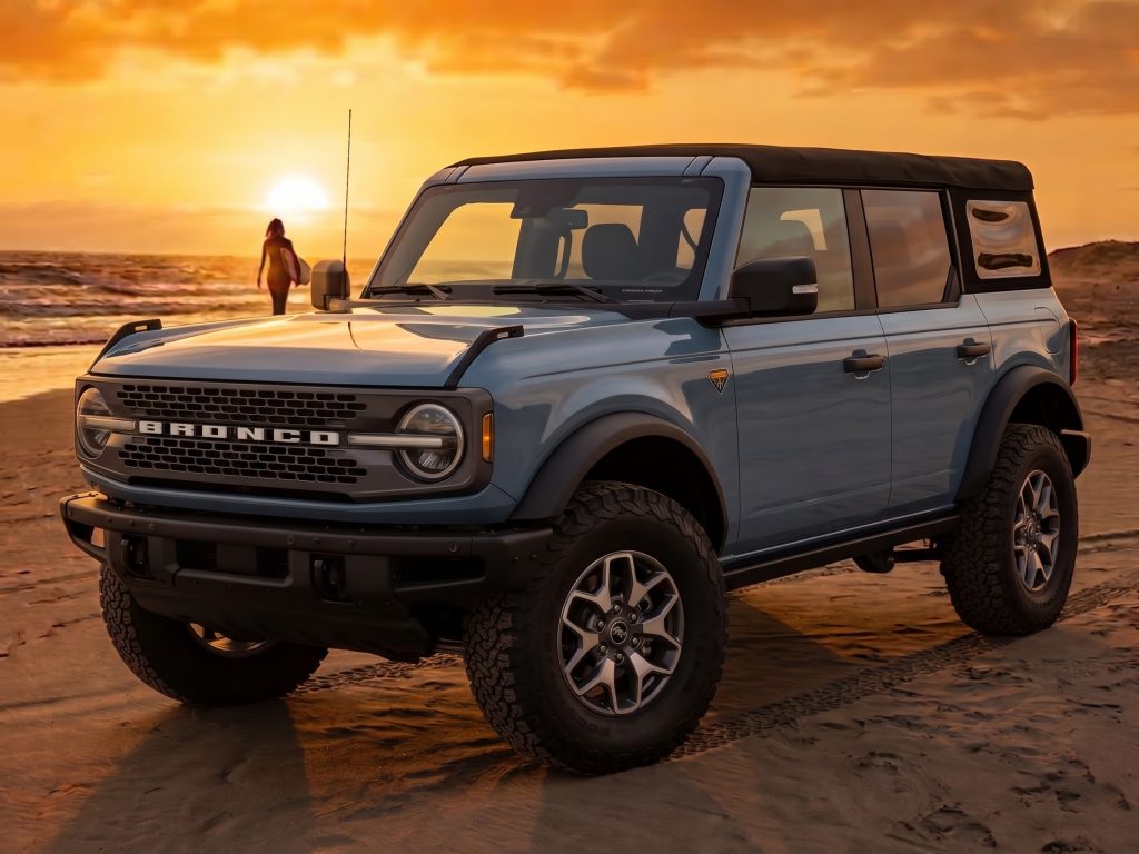 A light blue, four-door Ford Bronco Badlands edition parked on a sandy beach at sunset. In the background, a person carrying a surfboard walks toward the ocean waves under a vibrant orange sky."