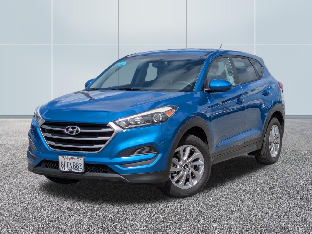 A three-quarter front view of a bright blue Hyundai Tucson (third generation, circa 2016–2018) parked on a grey asphalt surface against a clean, white paneled wall. The vehicle features: A prominent hexagonal chrome grille with the Hyundai logo. Sleek, swept-back headlights and black plastic cladding along the wheel arches and side skirts. Multi-spoke silver alloy wheels. A California license plate reading "8FCV882" with a "Galpin Honda" dealer frame. The lighting is bright and even, highlighting the car's metallic finish and contoured side body lines.