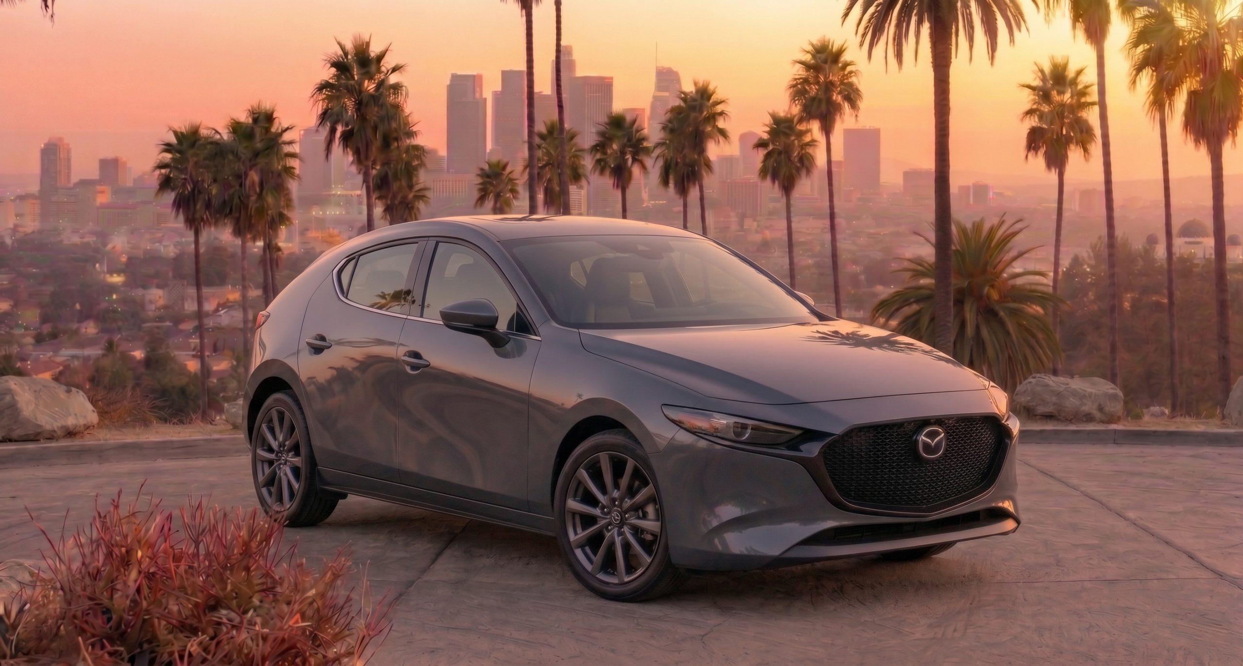 A grey Mazda3 hatchback, viewed from a front three-quarter angle, is parked on a concrete overlook at golden hour. Tall palm trees frame the car, with a hazy downtown Los Angeles skyline visible in the distance under a warm, pink-orange gradient sky. Foreground details include a reddish scrub bush and a large rock.