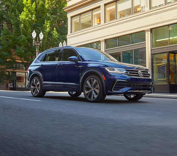 A Tiguan shown in Atlantic Blue Metallic drives through a modern, downtown area.