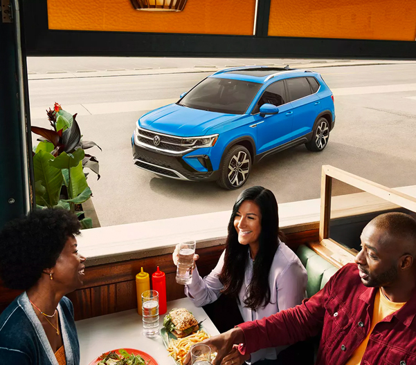 Three people seated in a booth at a diner, as the front driver side of a Taos in Cornflower Blue is seen through the window.