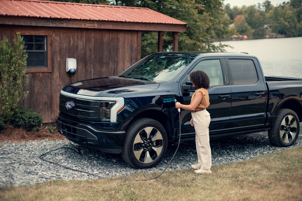 A woman charging her Ford F-150 Lightning using the Ford Connected Charge Station attached to her cabin in the background.